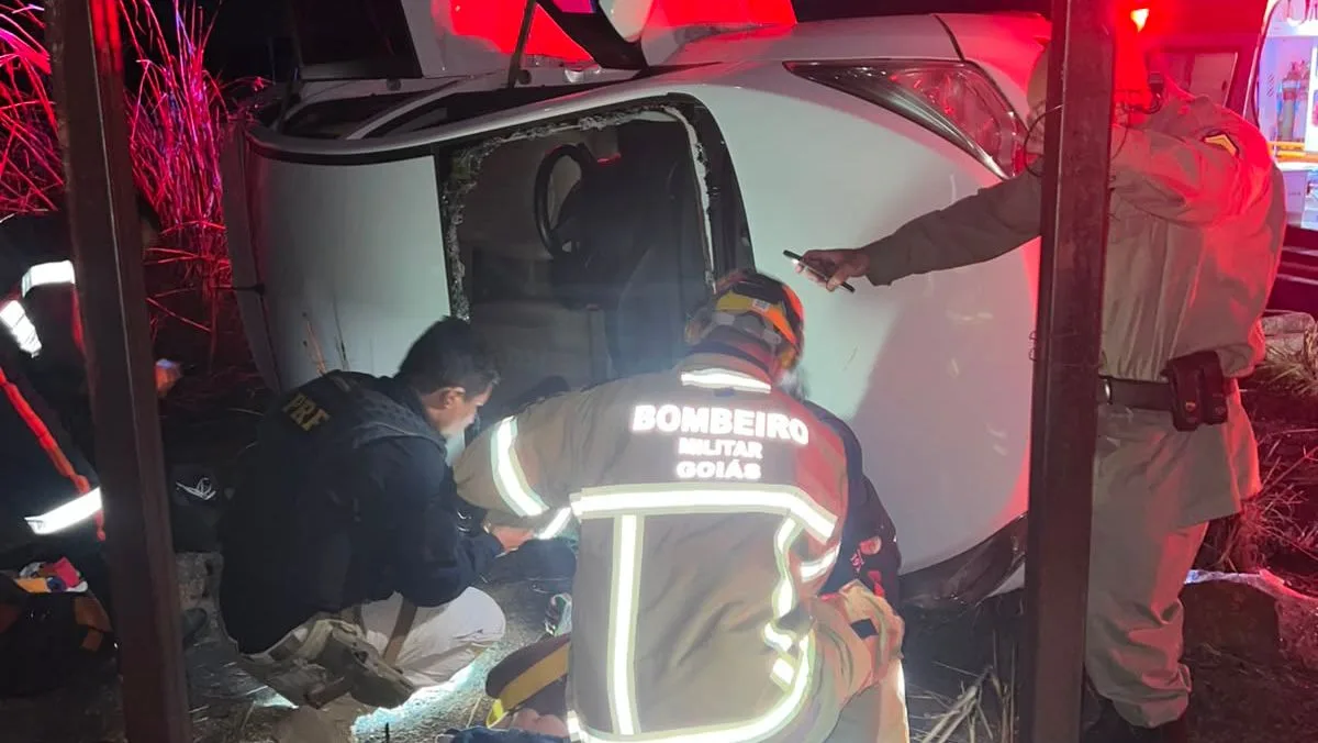 O close mostra o teto do carro amassado. O bombeiro, agachado e focado, remove detritos. Ele está vestido com um uniforme de segurança, incluindo um capacete e óculos de proteção. As peças do carro estão espalhadas ao redor dele, criando uma atmosfera de caos e perigo.