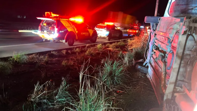 Esta imagem captura a cena do acidente em um ângulo amplo. O carro está de cabeça para baixo e os bombeiros estão trabalhando duro para libertar o motorista preso. O teto amassado do carro é visível e o homem em perigo é visto pendurado dentro. Os bombeiros estão usando ferramentas de corte e outros equipamentos para abrir o carro e salvar o motorista.