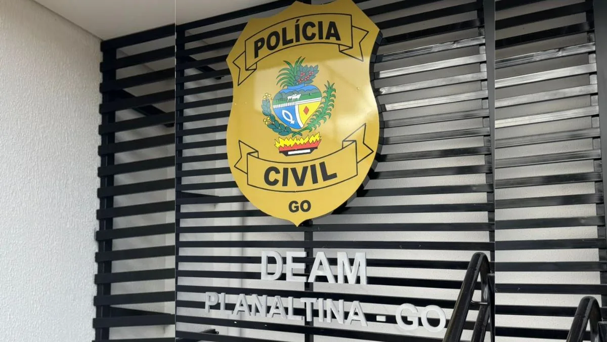 Uma imagem em close de um brasão da Polícia Civil de Goiás, em formato de escudo amarelo, com o brasão de armas central e o texto "POLÍCIA CIVIL" e "GO" em preto. O brasão está montado sobre uma parede de ripas pretas de alumínio. Abaixo do brasão, fixadas à mesma parede, estão as palavras "DEAM" e, na linha inferior, "PLANALTINA - GO", em letras brancas, maiúsculas e metálicas, de forma clara e visível. A iluminação é difusa.