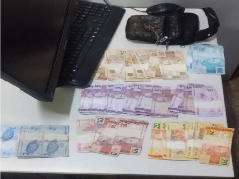 Dinheiro recuperado pela Polícia Militar após um roubo é exibido sobre uma mesa branca, ao lado de uma pochete escura, monitor de computador, teclado e mouse. As cédulas de diferentes valores estão organizadas e totalizam o montante apreendido durante a ação policial em Formosa.