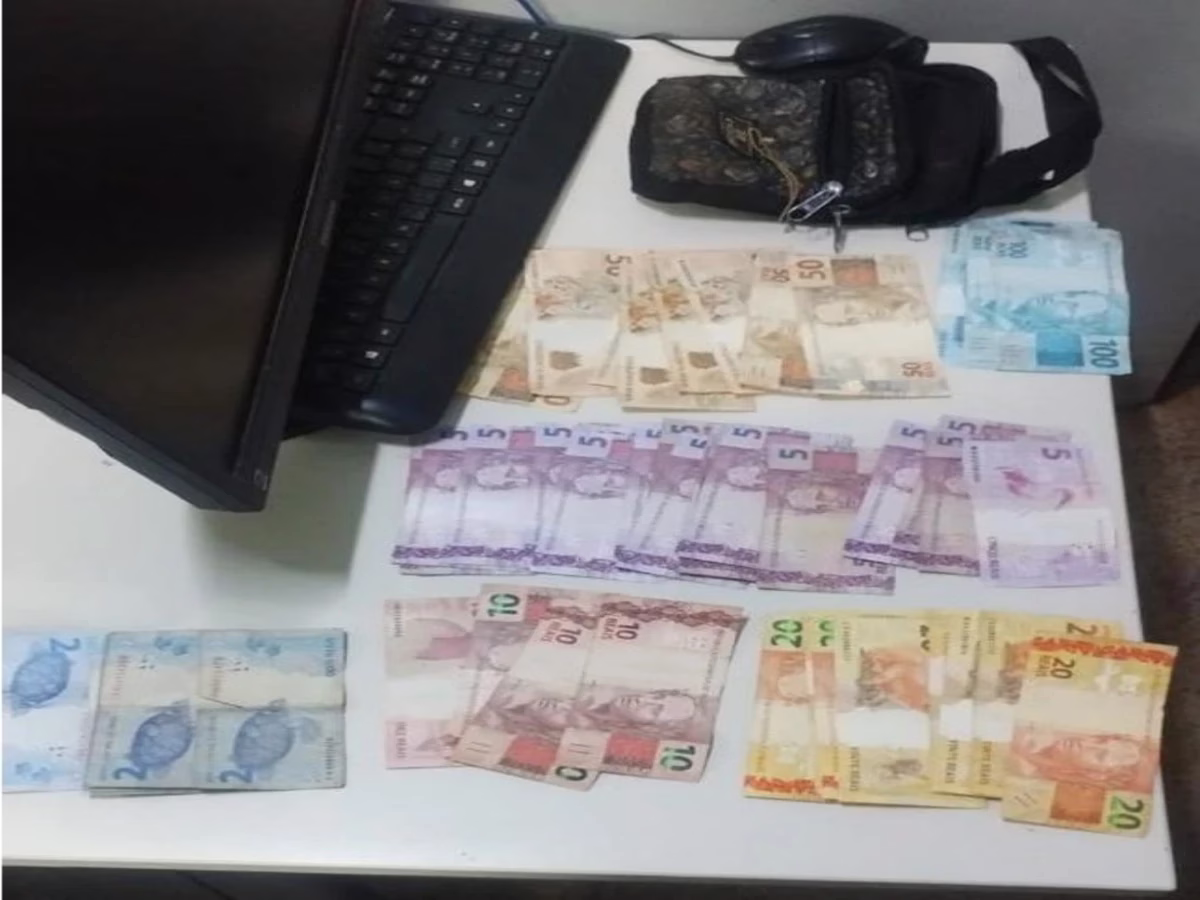 Dinheiro recuperado pela Polícia Militar após um roubo é exibido sobre uma mesa branca, ao lado de uma pochete escura, monitor de computador, teclado e mouse. As cédulas de diferentes valores estão organizadas e totalizam o montante apreendido durante a ação policial em Formosa.