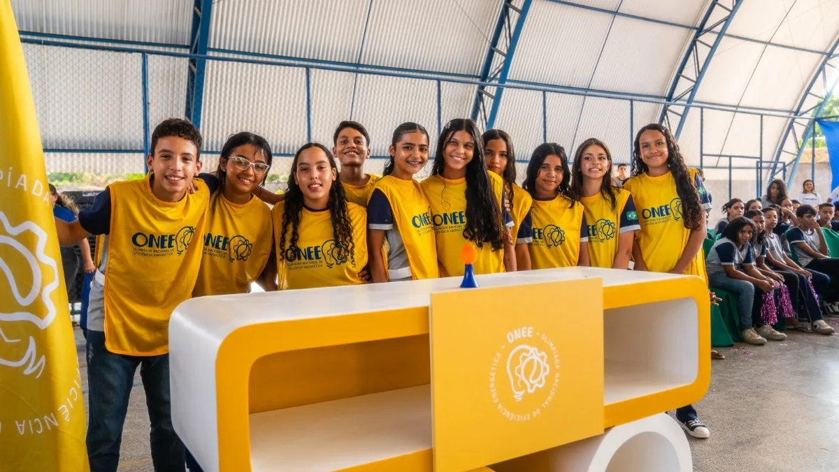 Grupo de estudantes usando camisetas amarelas na Olimpíada Nacional de Eficiência Energética (ONEE) posa para uma foto em um ginásio escolar, atrás de um estande com a identidade visual do evento e uma bandeira da competição, durante uma atividade educativa sobre eficiência energética.