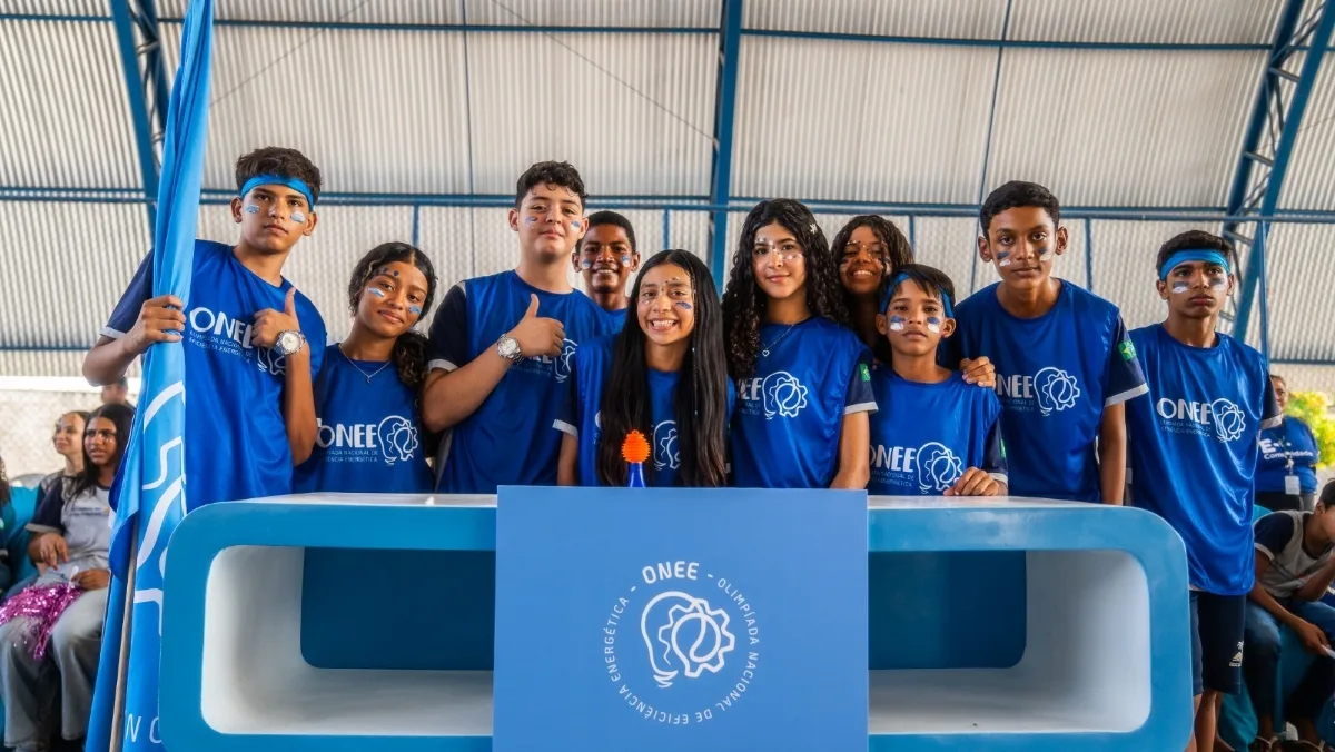 Grupo de estudantes usando camisetas azuis participando da Olimpíada Nacional de Eficiência Energética (ONEE) posa para uma foto em um ginásio escolar, atrás de um estande com a identidade visual do evento e uma bandeira da competição, durante uma atividade educativa sobre eficiência energética.