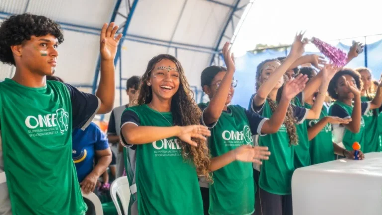 Estudantes usando camisetas verdes da Olimpíada Nacional de Eficiência Energética (ONEE) participam de uma atividade interativa em um ginásio, com as mãos levantadas, sorrindo e demonstrando entusiasmo durante o evento educacional.