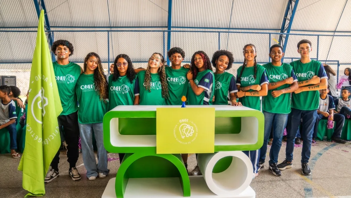 Grupo de estudantes usando camisetas verdes da Olimpíada Nacional de Eficiência Energética (ONEE) posa para uma foto em um ginásio escolar, atrás de um estande com a identidade visual do evento e uma bandeira da competição, durante uma atividade educativa sobre eficiência energética.