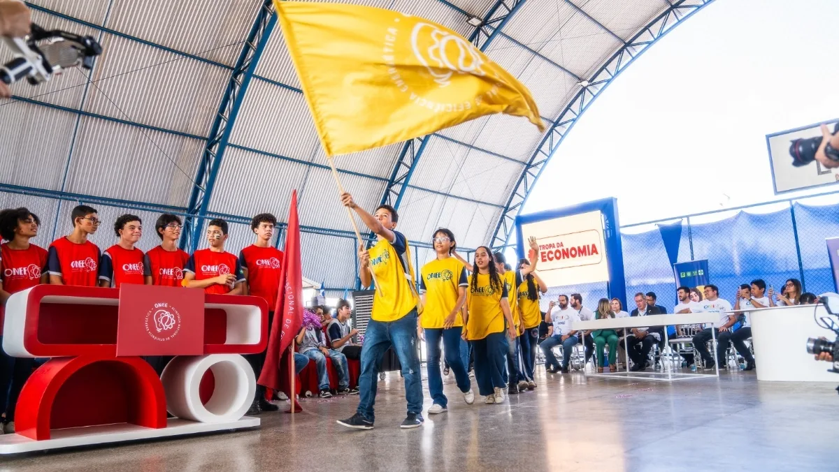 Grupo de estudantes usando camisetas vermelhas durante Olimpíada Nacional de Eficiência Energética (ONEE) posa para uma foto em um ginásio escolar, atrás de um estande com a identidade visual do evento e uma bandeira da competição, durante uma atividade educativa sobre eficiência energética.