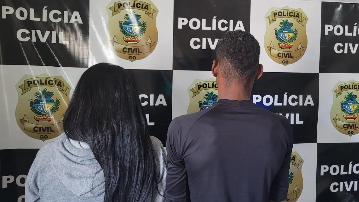 Uma mulher de longos cabelos pretos e jaqueta cinza e um homem de camisa preta posam de costas para a câmera contra uma parede quadriculada em preto e branco. ​A parede é composta por um padrão repetitivo: quadrados pretos com o texto "POLÍCIA CIVIL" e quadrados brancos com um brasão dourado que também diz "POLÍCIA CIVIL GO" (que significa Polícia Civil de Goiás). ​A imagem mostra os dois indivíduos sendo detidos e levados à delegacia.