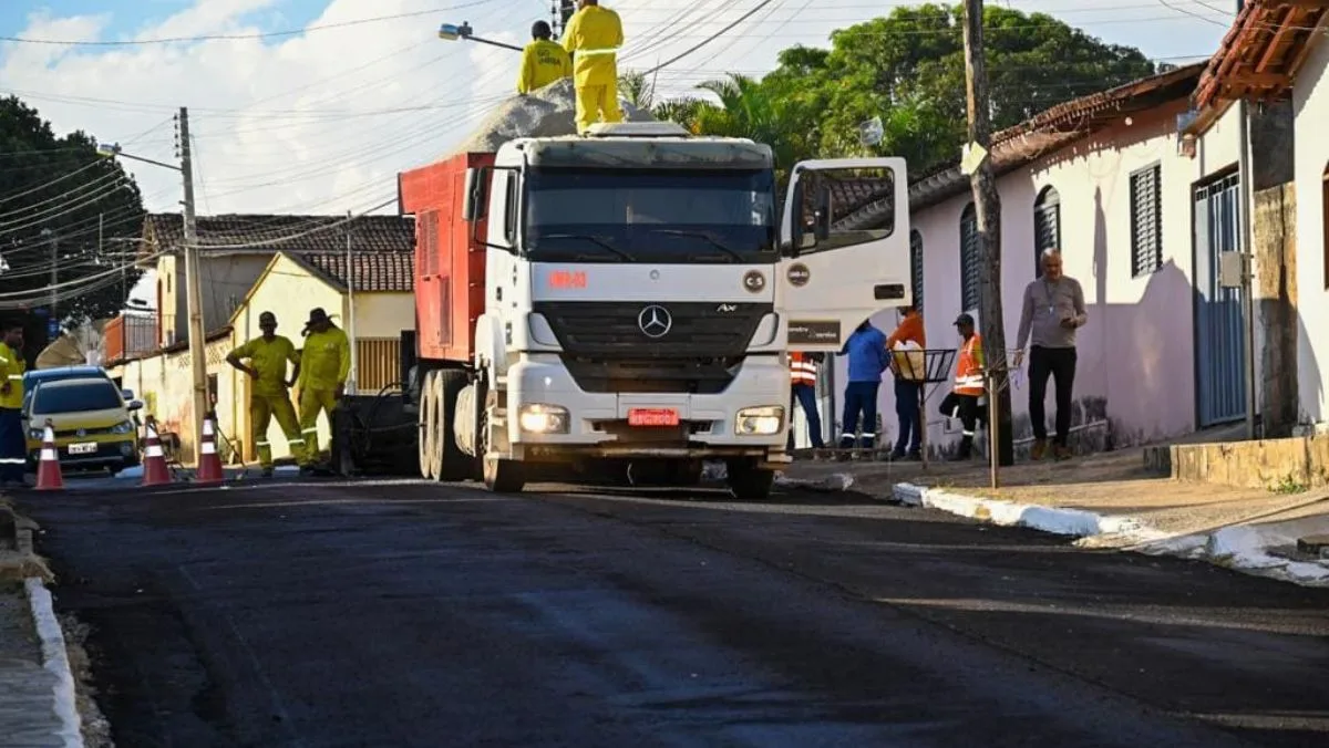 Caminhão, homens e máquinas trabalhando em recapeamento asfáltico.