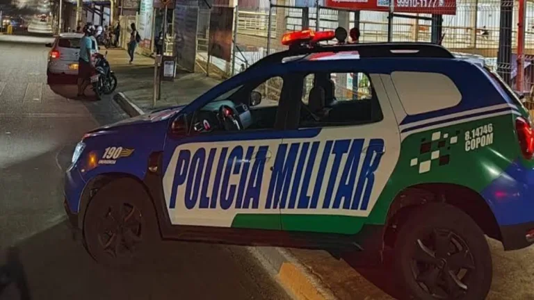 Viatura da Polícia Militar estacionada em uma avenida durante a noite, com giroflex ligado, em uma área urbana com veículos, motocicletas e estabelecimentos comerciais ao fundo.