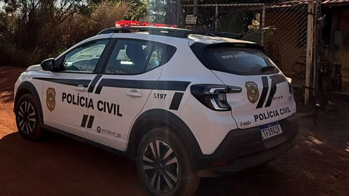 Uma fotografia externa e diurna de uma viatura da Polícia Civil de Goiás, um SUV branco (marca Renault Duster), estacionado em uma estrada de terra. A viatura está de traseira e lateral, mostrando a porta traseira com o emblema da Polícia Civil, o texto "POLÍCIA CIVIL" e a inscrição "197". A placa traseira, legível, é "TF09A19". O veículo está em frente a uma casa de campo com um muro de arame e vegetação densa. O sol brilha forte, criando sombras, e há árvores e uma cerca ao fundo. As luzes de emergência (giroflex) no teto estão desligadas.