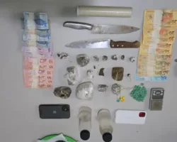 Vista superior de diversos itens apreendidos dispostos sobre uma mesa cinza. Há cédulas de Real brasileiro organizadas em fileiras nas laterais esquerda e direita, duas facas grandes de cozinha, um rolo de plástico filme, um distintivo metálico da Polícia Civil, dois smartphones (um preto e um branco), duas pequenas garrafas plásticas transparentes com tampas pretas, uma mini balança digital de precisão e dezenas de porções de entorpecentes, incluindo tabletes e trouxinhas de maconha, além de pequenos comprimidos verdes e amarelos (supostamente ecstasy) enfileirados.