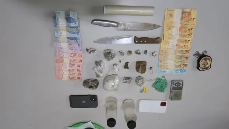 Vista superior de diversos itens apreendidos dispostos sobre uma mesa cinza. Há cédulas de Real brasileiro organizadas em fileiras nas laterais esquerda e direita, duas facas grandes de cozinha, um rolo de plástico filme, um distintivo metálico da Polícia Civil, dois smartphones (um preto e um branco), duas pequenas garrafas plásticas transparentes com tampas pretas, uma mini balança digital de precisão e dezenas de porções de entorpecentes, incluindo tabletes e trouxinhas de maconha, além de pequenos comprimidos verdes e amarelos (supostamente ecstasy) enfileirados.