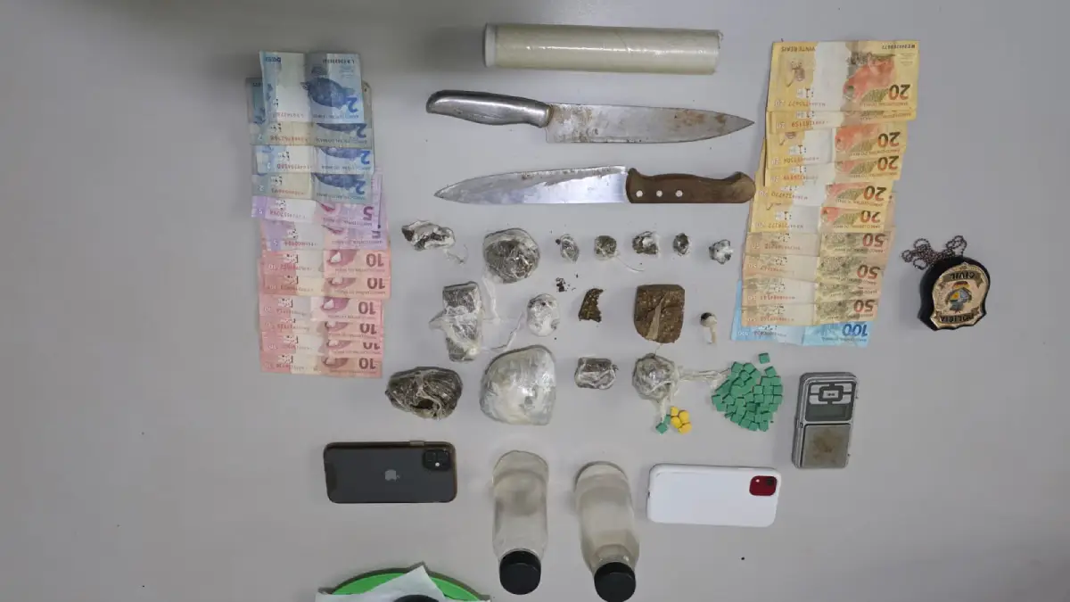 Vista superior de diversos itens apreendidos dispostos sobre uma mesa cinza. Há cédulas de Real brasileiro organizadas em fileiras nas laterais esquerda e direita, duas facas grandes de cozinha, um rolo de plástico filme, um distintivo metálico da Polícia Civil, dois smartphones (um preto e um branco), duas pequenas garrafas plásticas transparentes com tampas pretas, uma mini balança digital de precisão e dezenas de porções de entorpecentes, incluindo tabletes e trouxinhas de maconha, além de pequenos comprimidos verdes e amarelos (supostamente ecstasy) enfileirados.