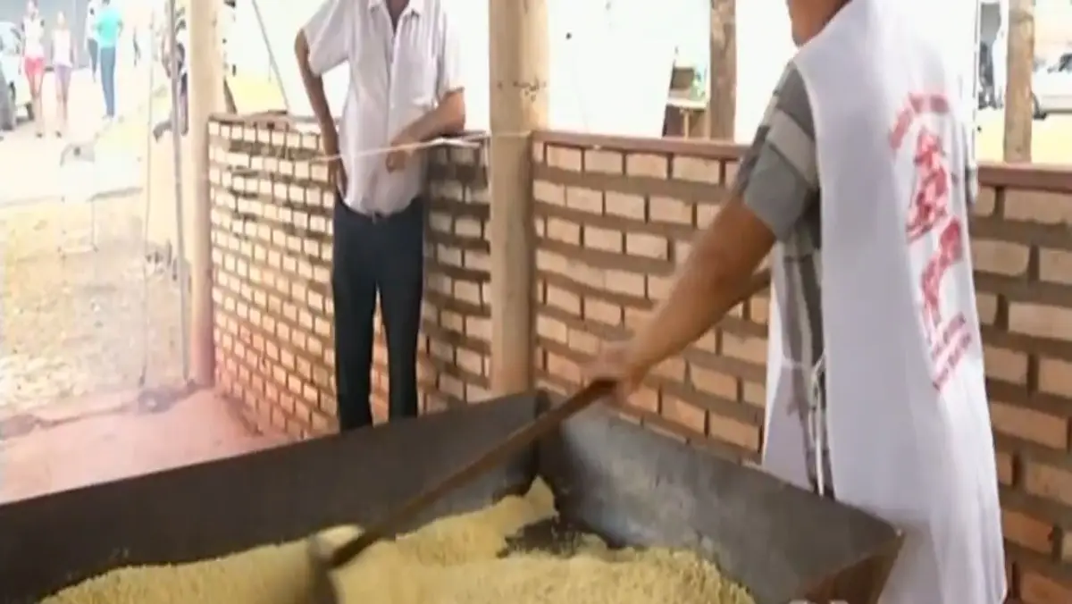 Uma pessoa de costas usando um colete branco mexe uma grande quantidade de farinha amarela em uma moenda ou tacho retangular longo de metal com um rodo de madeira. Ao fundo, um homem com camisa branca observa o processo encostado em uma pilastra de tijolos.