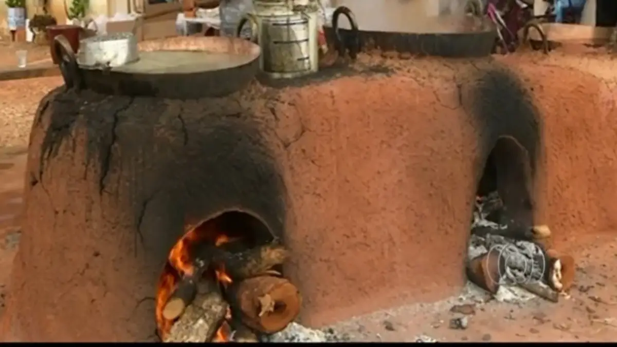 A tradição está de volta!
Um grande fogão a lenha tradicional feito de barro avermelhado com duas aberturas para o fogo. Na abertura da esquerda, há chamas ativas consumindo lenhas grossas. No topo do fogão, descansam tachos e panelas grandes de metal usados para a moagem.
