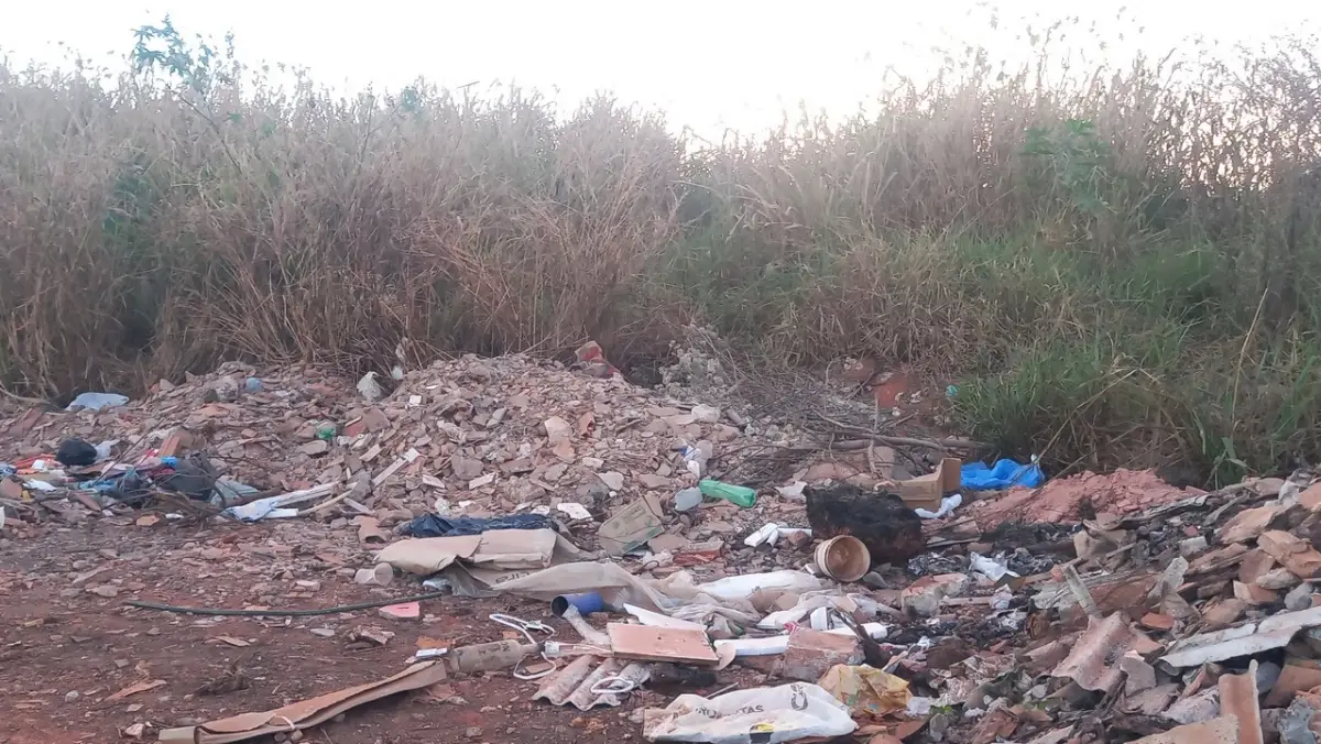 Grande acúmulo de lixo e entulho de construção descartados irregularmente junto a um matagal alto. Entre os resíduos visíveis estão pedaços de tijolos, telhas, sacolas plásticas e garrafas descartáveis.
