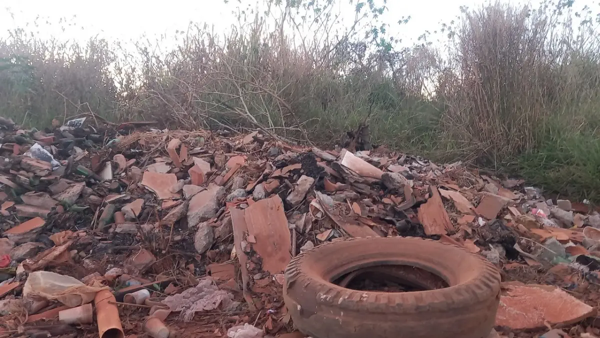 Depósito de lixo e entulho a céu aberto com um pneu de caminhão velho em destaque no primeiro plano. Ao fundo, uma grande pilha de restos de telhas, tijolos e plásticos acumulados ao lado de vegetação seca.