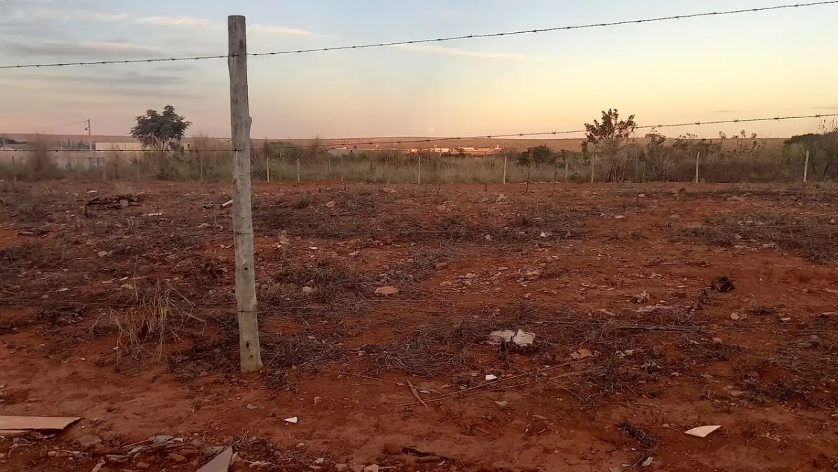 Terreno baldio amplo com terra batida avermelhada e vegetação rasteira seca. O local é cercado por estacas de madeira e arame farpado, com algumas árvores ao fundo sob um céu claro de fim de tarde.
