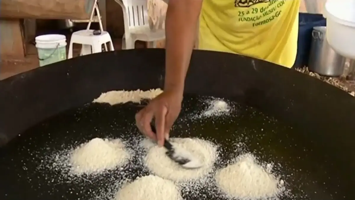 Close-up da mão de uma pessoa moldando pequenas porções de massa branca ou polvilho dentro de uma grande chapa de metal escura e arredondada. Ao fundo, uma pessoa veste uma camiseta amarela com os dizeres "Fundação Museu Couros".