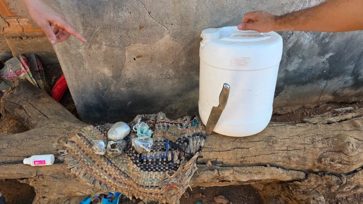 Imagem em plano detalhe mostrando objetos sobre um tronco de árvore caído em ambiente externo. No centro, há um galão plástico branco de formato cilíndrico com uma faca de cozinha presa verticalmente em sua lateral; uma mão segura a alça superior do galão. À esquerda, sobre um pedaço de pano xadrez velho e desfiado, estão espalhadas pequenas porções de substâncias embaladas em sacos plásticos transparentes. No canto esquerdo, sobre o tronco, há um frasco cilíndrico branco com rótulo rosa. No canto superior esquerdo, outra mão aponta em direção aos objetos. Ao fundo, vê-se uma parede de concreto rústica e desgastada.