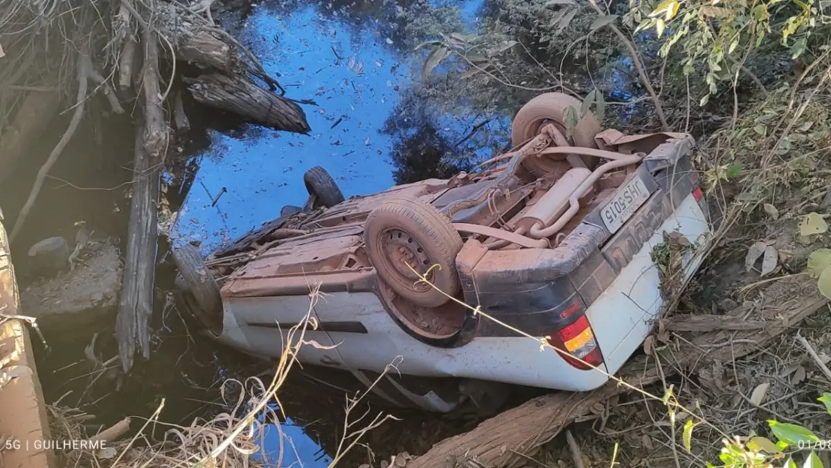 Carro caído em corrego após sai sa pista, na imagem é possível ver o carro com as rodas pra cima, corrego com água e vegetação.